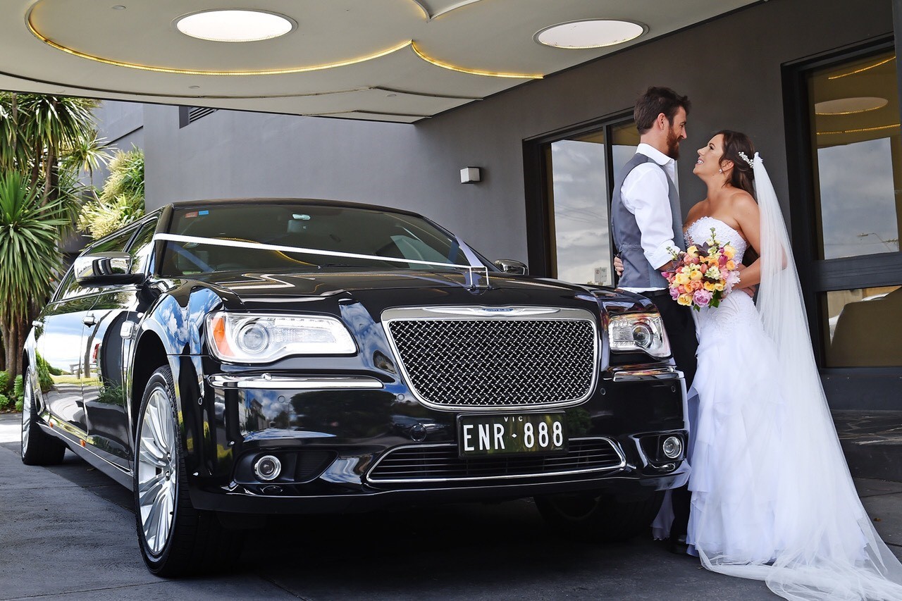 Bride and groom standing beside a black stretch limousine at a wedding venue.
